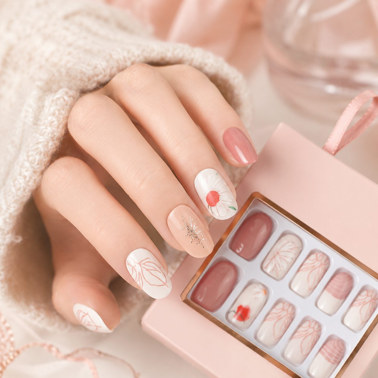 Light skin tone hand wearing glossy floral press on nails with pink nude designs, shown in a close-up lifestyle manicure photo with product packaging.