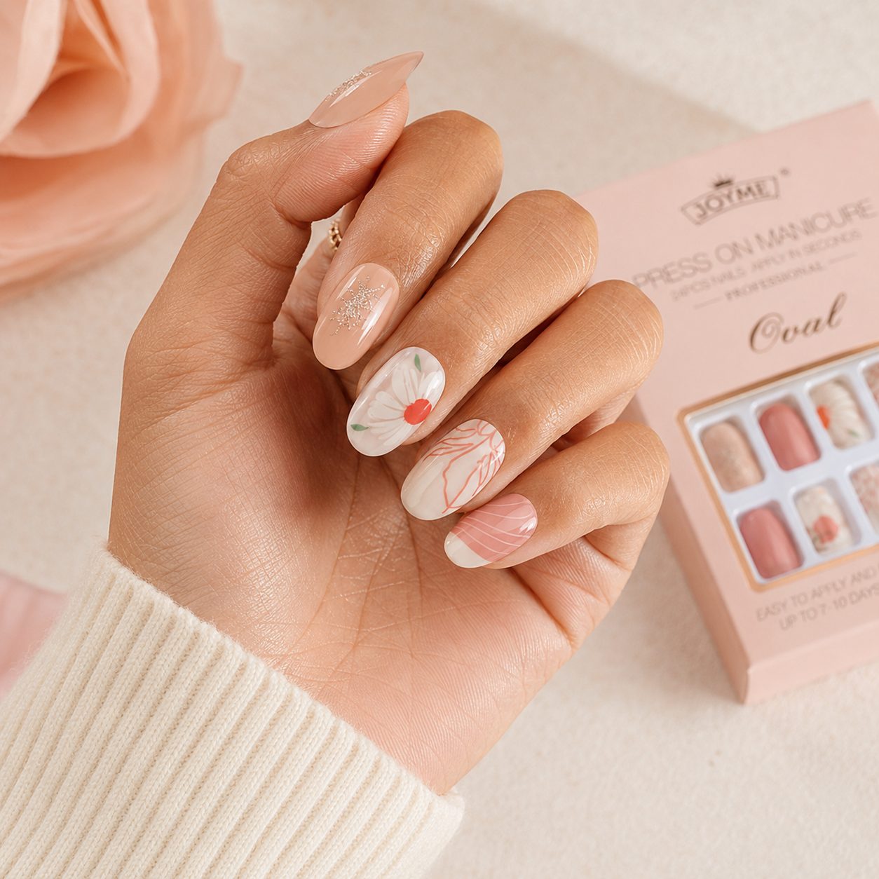 Medium skin tone hand wearing nude floral press on nails with glossy finish, featuring a realistic close-up manicure look beside the product box.